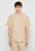 Junger Mann mit kurzen braunen Haaren, der ein beiges, kurzärmeliges, strukturiertes T-Shirt und passende beige Hosen trägt, steht vor einem schlichten hellen Hintergrund.