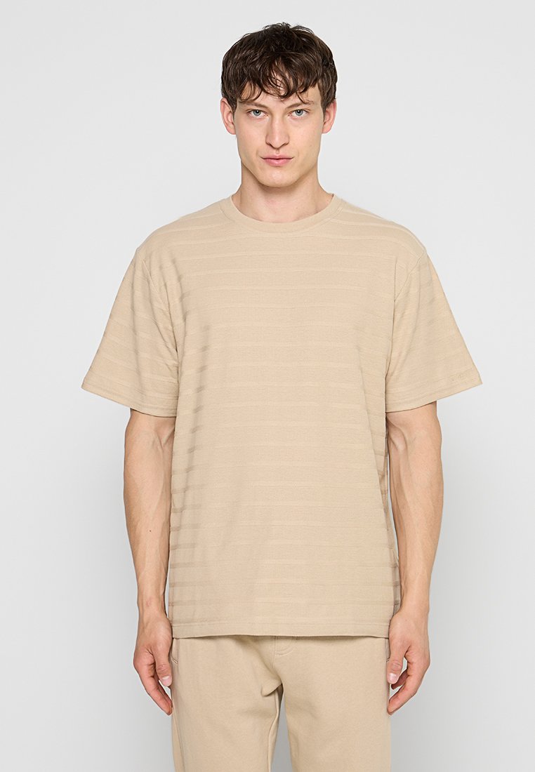Junger Mann mit kurzen braunen Haaren, der ein beiges, kurzärmeliges, strukturiertes T-Shirt und passende beige Hosen trägt, steht vor einem schlichten hellen Hintergrund.