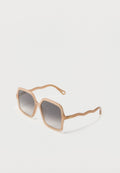 Quadratisch geformte Sonnenbrille mit beigen Rahmen, verlaufend dunklen Gläsern und welligen beigen Bügeln, die auf einer weißen Oberfläche liegen.