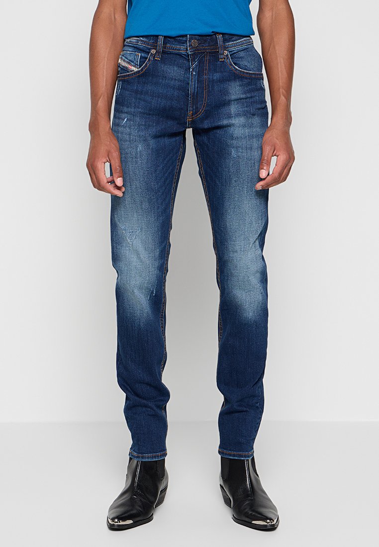 Dunkelblaue Jeans im Slim-Fit-Stil mit ausgebleichten Stellen, gestickten Akzenten und einem Fünf-Taschen-Design. Kombiniert mit schwarzen Knöchelstiefeln.