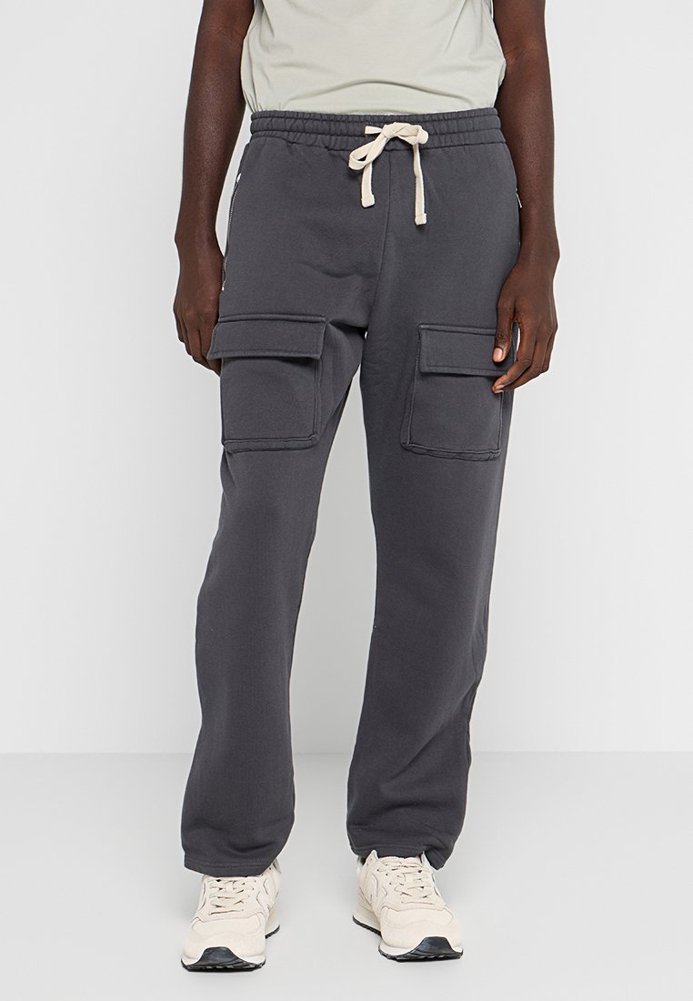 Person in dunkelgrauen Cargo-Sweatpants mit Kordelzug und beigen Sneakers vor einem schlichten Hintergrund.