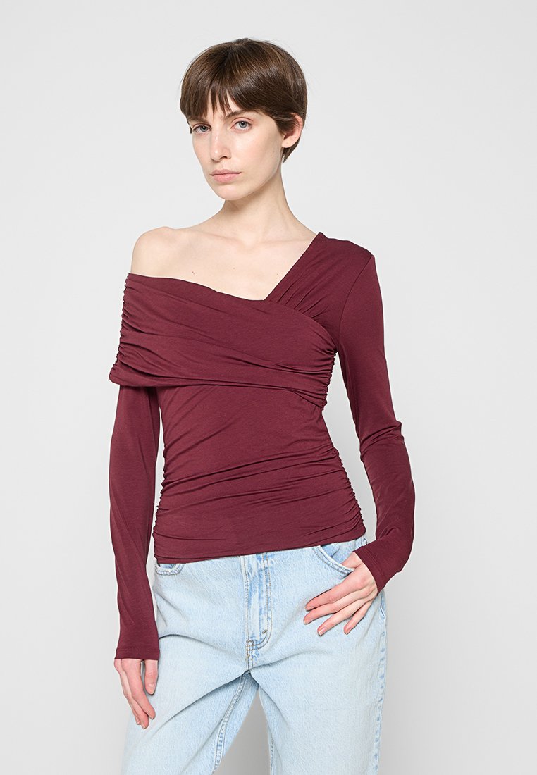 Junge Frau mit kurzen braunen Haaren, die ein burgunderfarbenes asymmetrisches Schulterfreies Oberteil und hellblaue hoch taillierte Jeans trägt und vor einem schlichten Hintergrund steht.