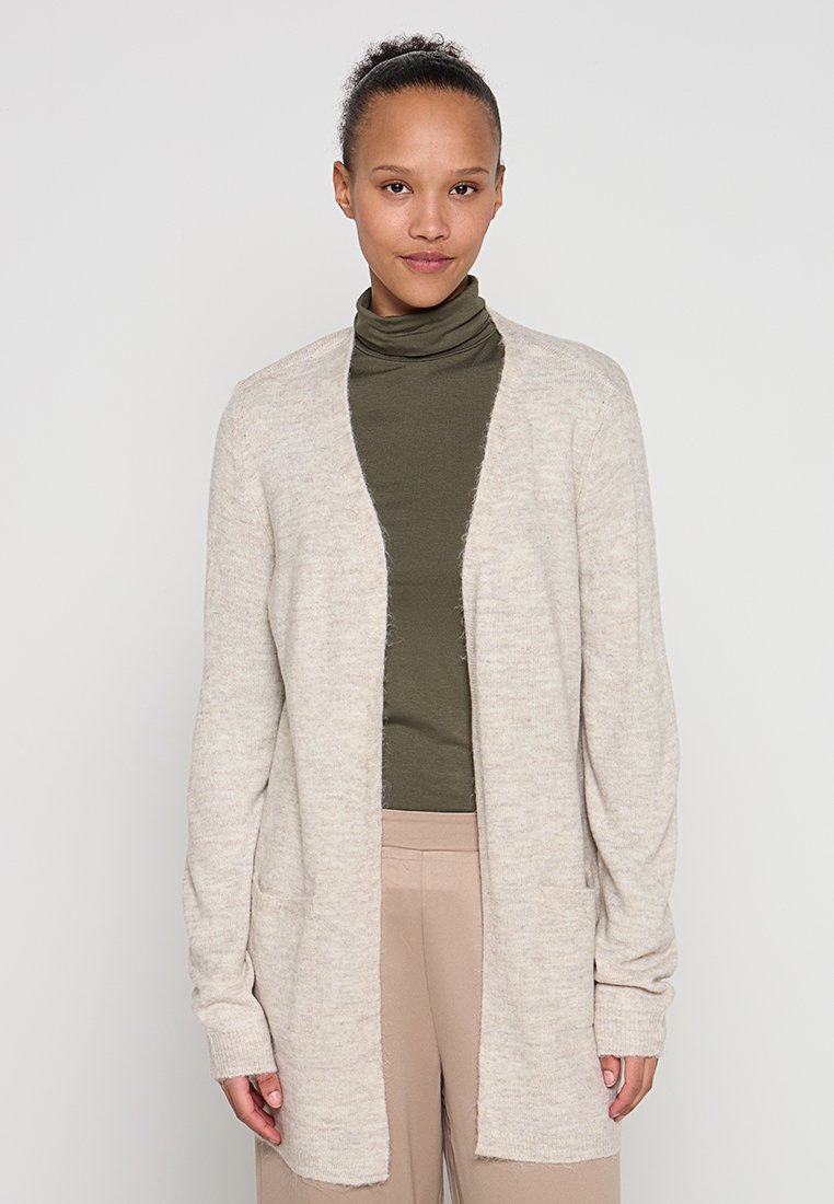 Beiger Strickjacke mit offenem Vorderteil, langen Ärmeln und Seitentaschen. Weicher, strukturierter Strickstoff. Getragen über einem dunkelgrünen Rollkragenpullover.