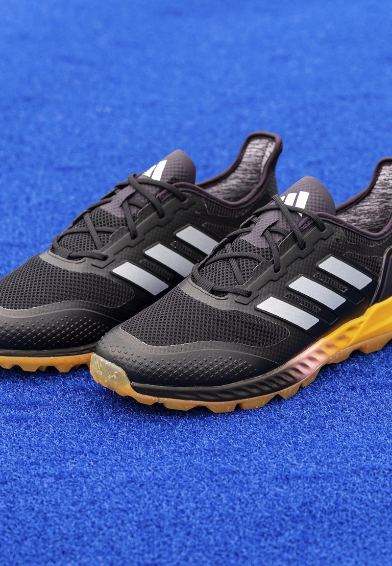 Schwarze Sportschuhe mit Mesh-Obermaterial, drei weißen Streifen, Gummisohle in Orange und Schwarz. Verfügt über strukturierte Muster und Schnürsenkel.