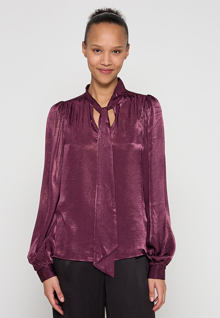 Lila Satinbluse mit einer Bindeschleife am Ausschnitt, langen Puffärmeln und gerafften Schultern. Glatte Textur und dezenter Glanz.