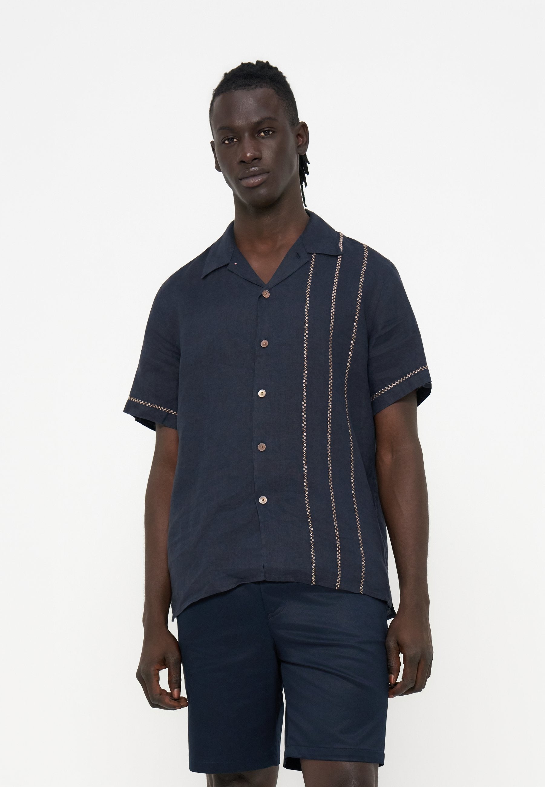 Mann mit dunkler Haut und kurzen Dreadlocks, der ein marineblaues kurzärmeliges Hemd mit vertikalen Nähten und passende marineblaue Shorts trägt.