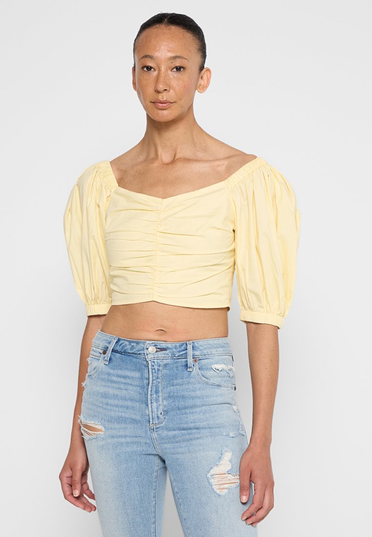 Gelbes cropped Oberteil mit kurzen Puffärmeln und einem gerafften Frontdesign. Kombiniert mit hellblauen, distressed Jeans.