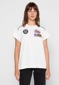 Weiße Baumwoll-T-Shirt mit kurzen Ärmeln, ausgestattet mit gestickten Applikationen: Schachbrettmuster, Blumenmuster und mehrfarbige Designs. Schwarze Akzente an den Schultern.