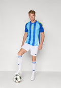 Blau- und hellblau gestreiftes Fußballtrikot mit gelben Akzenten, weiße Shorts und weiße Socken, steht auf einem Fußball in einer konzentrierten Haltung.
