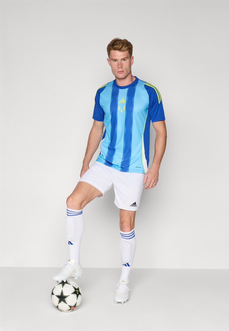 Blau- und hellblau gestreiftes Fußballtrikot mit gelben Akzenten, weiße Shorts und weiße Socken, steht auf einem Fußball in einer konzentrierten Haltung.