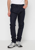 Mann in dunkelblauen Jeans und silber-grauen Sportschuhen, der mit einer Hand in der Tasche vor einem schlichten hellen Hintergrund steht.
