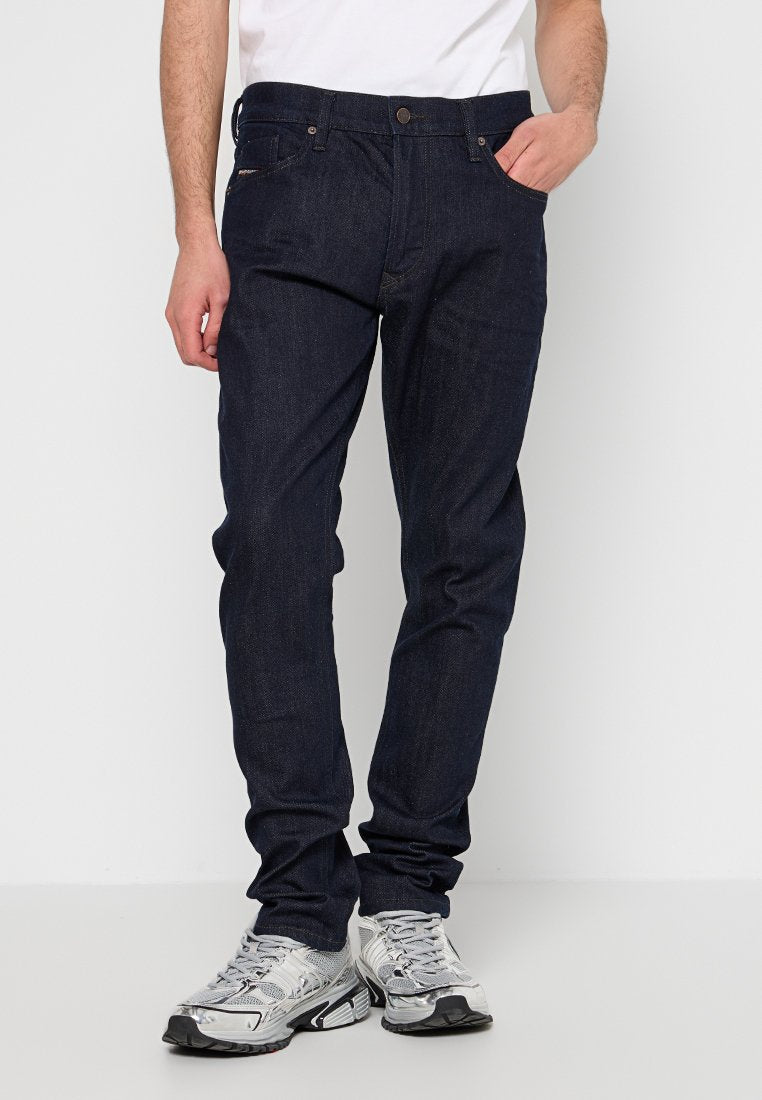 Mann in dunkelblauen Jeans und silber-grauen Sportschuhen, der mit einer Hand in der Tasche vor einem schlichten hellen Hintergrund steht.