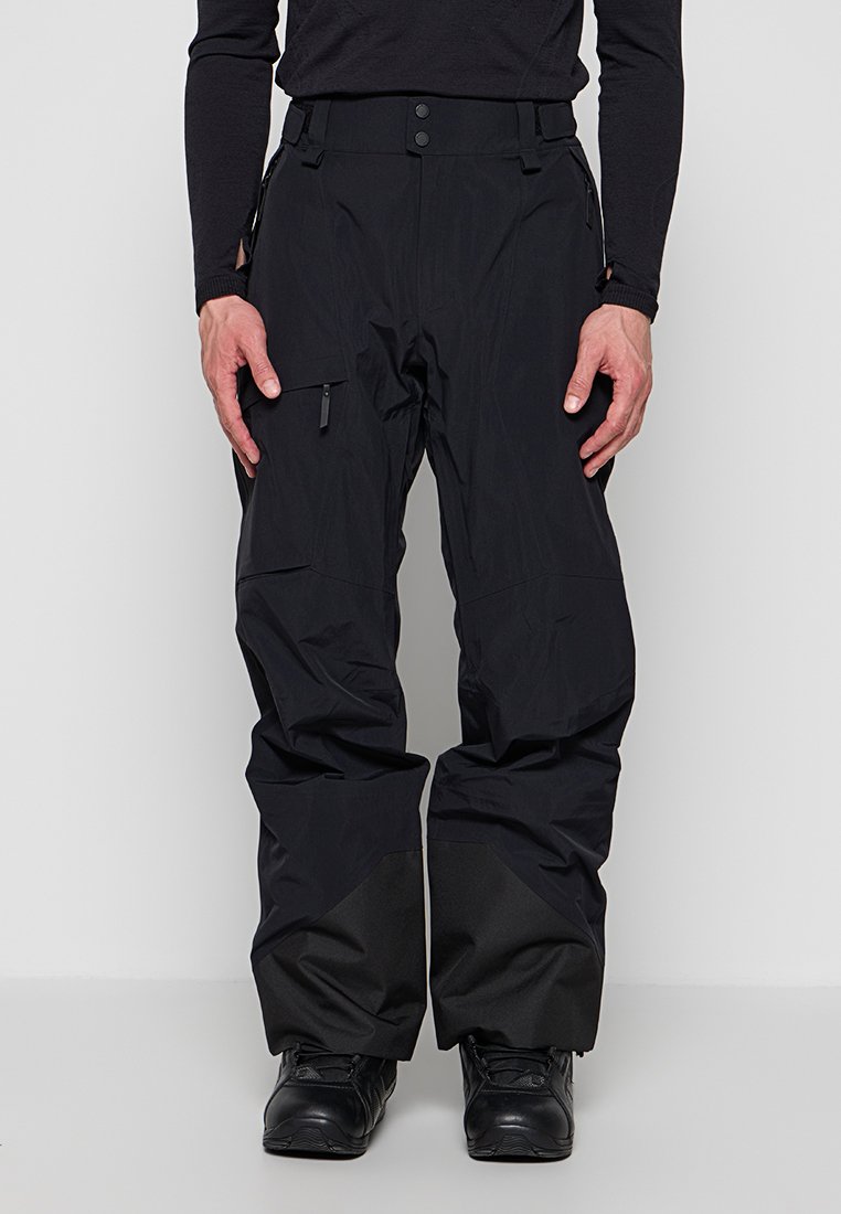 Mann trägt schwarze Schneehose mit Reißverschlusstasche am Oberschenkel und verstärktem Stoff an den unteren Beinen, kombiniert mit schwarzen Stiefeln und langärmligem Oberteil.