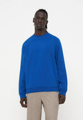 Mann mit dunkler Haut und geflochtenen Haaren, der einen hellblauen Pullover und hellbeige Hosen trägt, steht vor einem einfachen weißen Hintergrund.