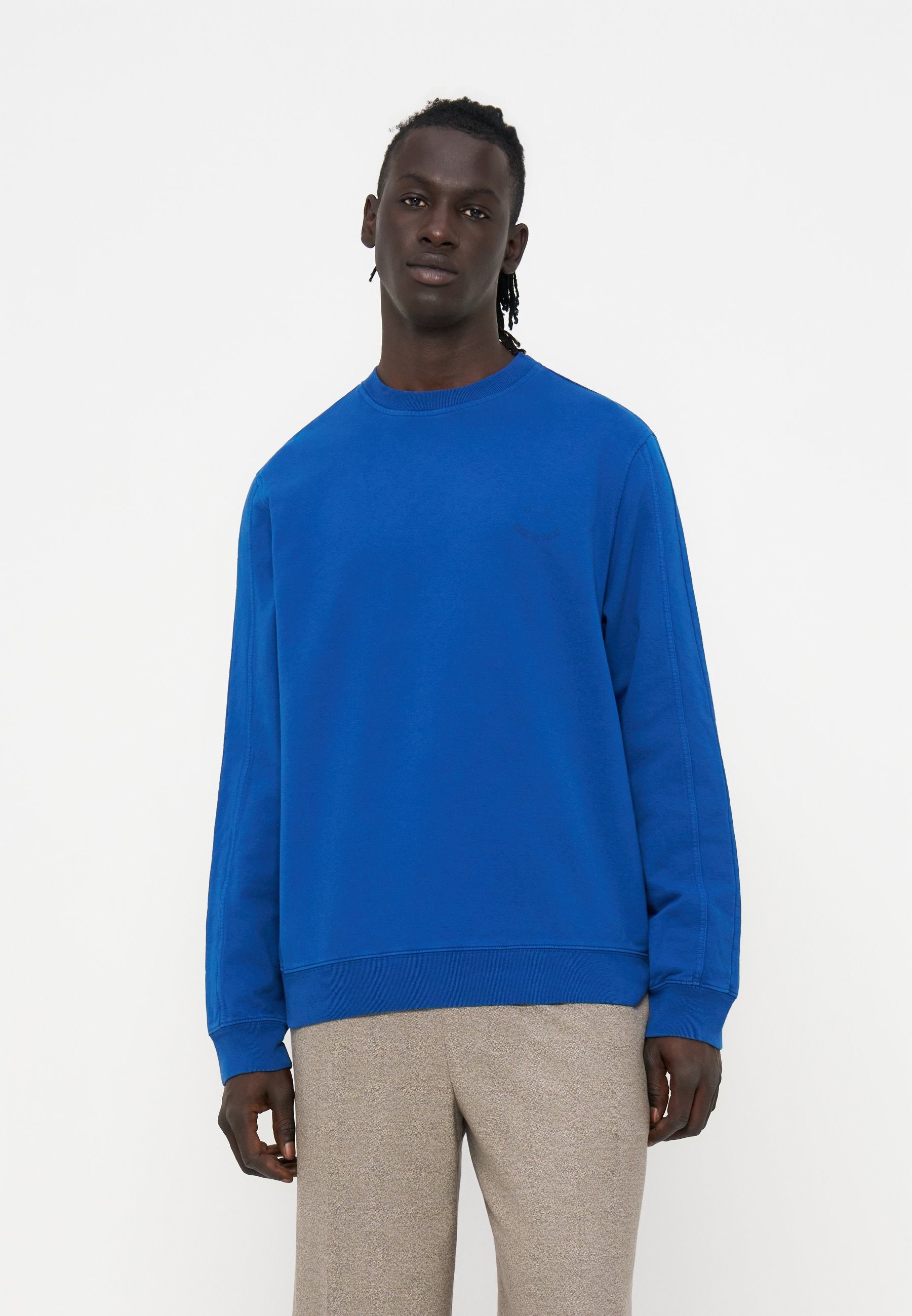 Mann mit dunkler Haut und geflochtenen Haaren, der einen hellblauen Pullover und hellbeige Hosen trägt, steht vor einem einfachen weißen Hintergrund.