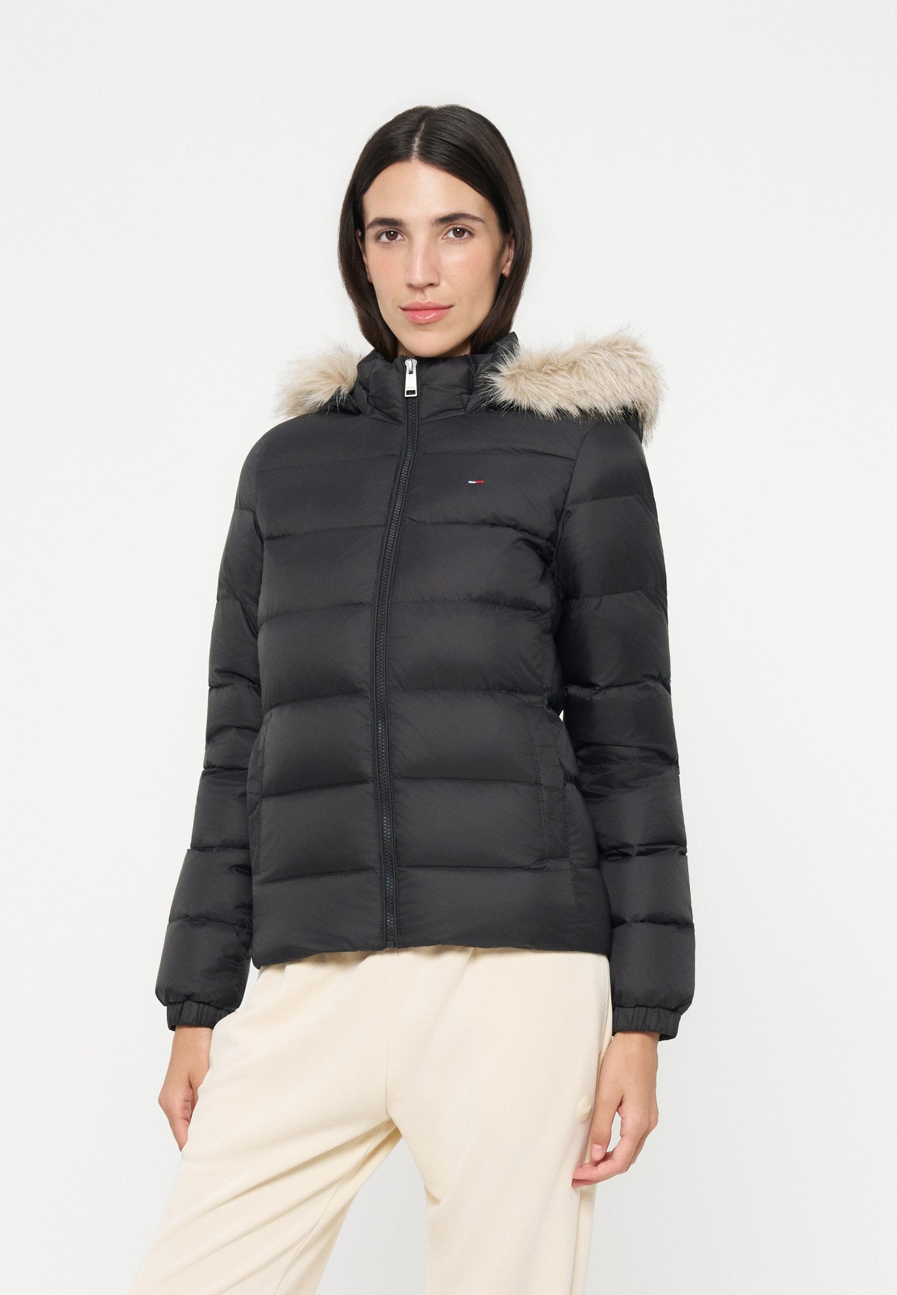 Frau trägt eine schwarze gesteppte Jacke mit pelzbesetzter Kapuze und beigen Hosen, steht vor einem weißen Hintergrund.