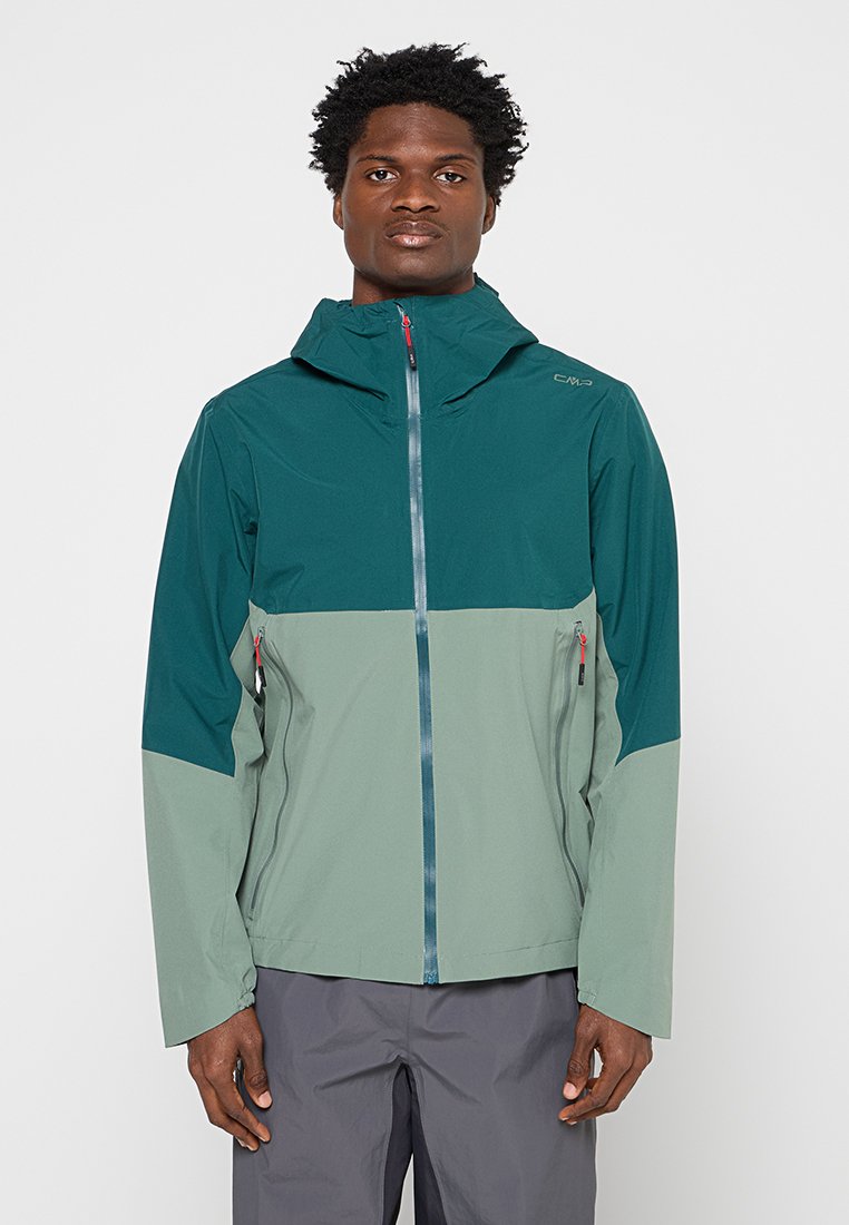 Mann steht nach vorne gerichtet, trägt eine türkis-hellgrüne Outdoorjacke mit Kapuzen und Reißverschlusstaschen sowie graue Hose.