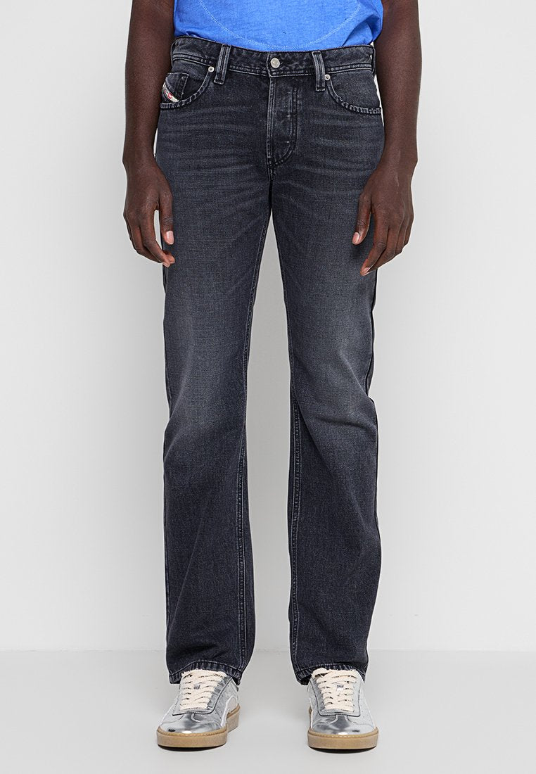 Person trägt ausgewaschene schwarze gerade Jeans, blauen Oberteil und silberne Sneakers, steht mit entspannten Händen an den Seiten.