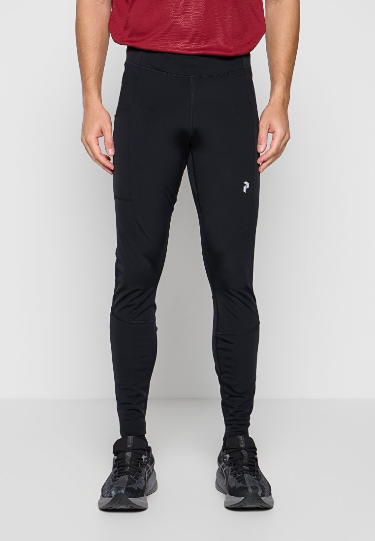 Mann trägt schwarze eng anliegende Sporthose mit Seitentasche, rotes strukturiertes Shirt und schwarze Laufschuhe, steht vor einfachem Hintergrund.