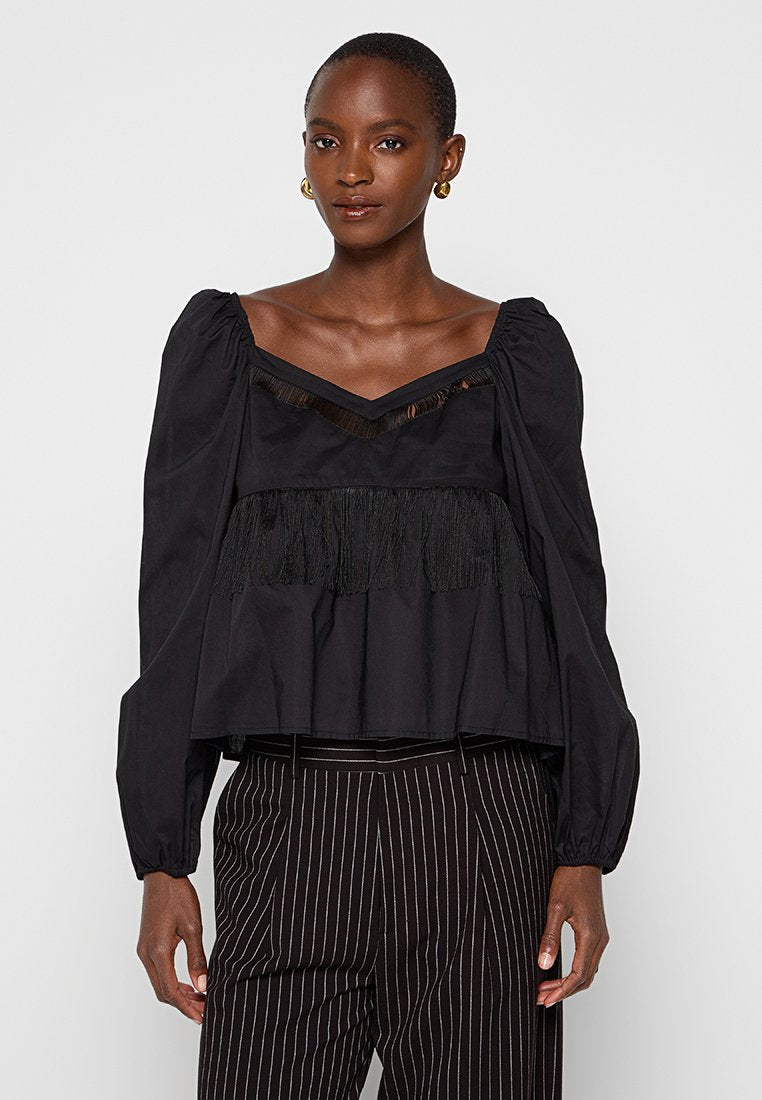 Schwarze Peplum-Bluse mit gepufften Ärmeln, strukturierten Fransen an dem Saum und einem V-Ausschnitt, aus glattem Stoff gefertigt. Kombiniert mit gestreiften Hosen.