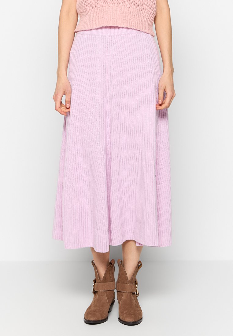 Frau in einem gerippten lavendelfarbenen Midiskirt, rosa figurbetontem ärmellosem Oberteil und braunen Wildleder-Ankle-Boots mit Schnallen.
