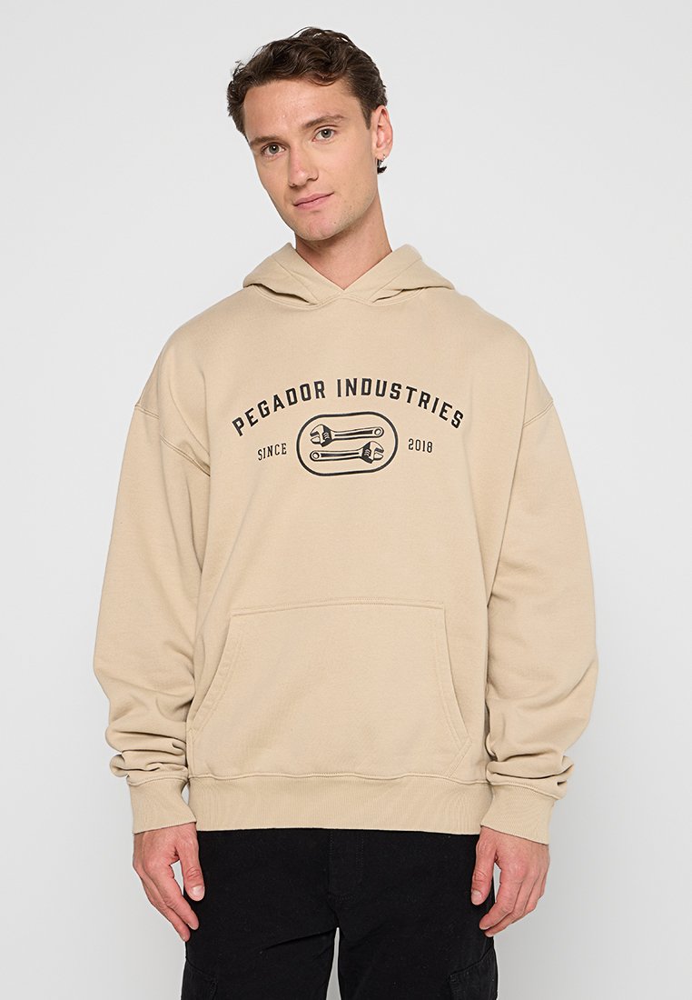 Beige Kapuzenpullover mit einer vorderen Kängurutasche, gerippten Bündchen und einem grafischen Textdesign, das "Pegador Industries" mit einem Schraubenschlüssel-Motiv zeigt.