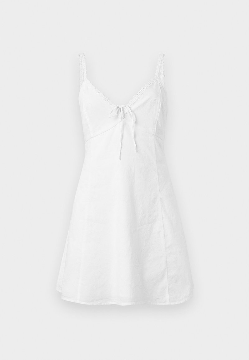 Weißes ärmelloses Sommerkleid mit dünnen Spitzenriemen und einer Schleifendetail vorne vor einfachem Hintergrund.