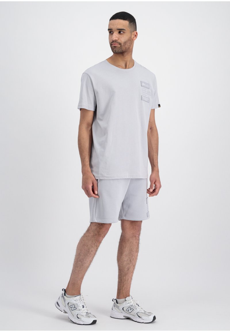 Helles graues T-Shirt mit kurzen Ärmeln, das drei strukturierte Taschen-Designs aufweist, kombiniert mit passenden hellgrauen Shorts und weißen Turnschuhen.