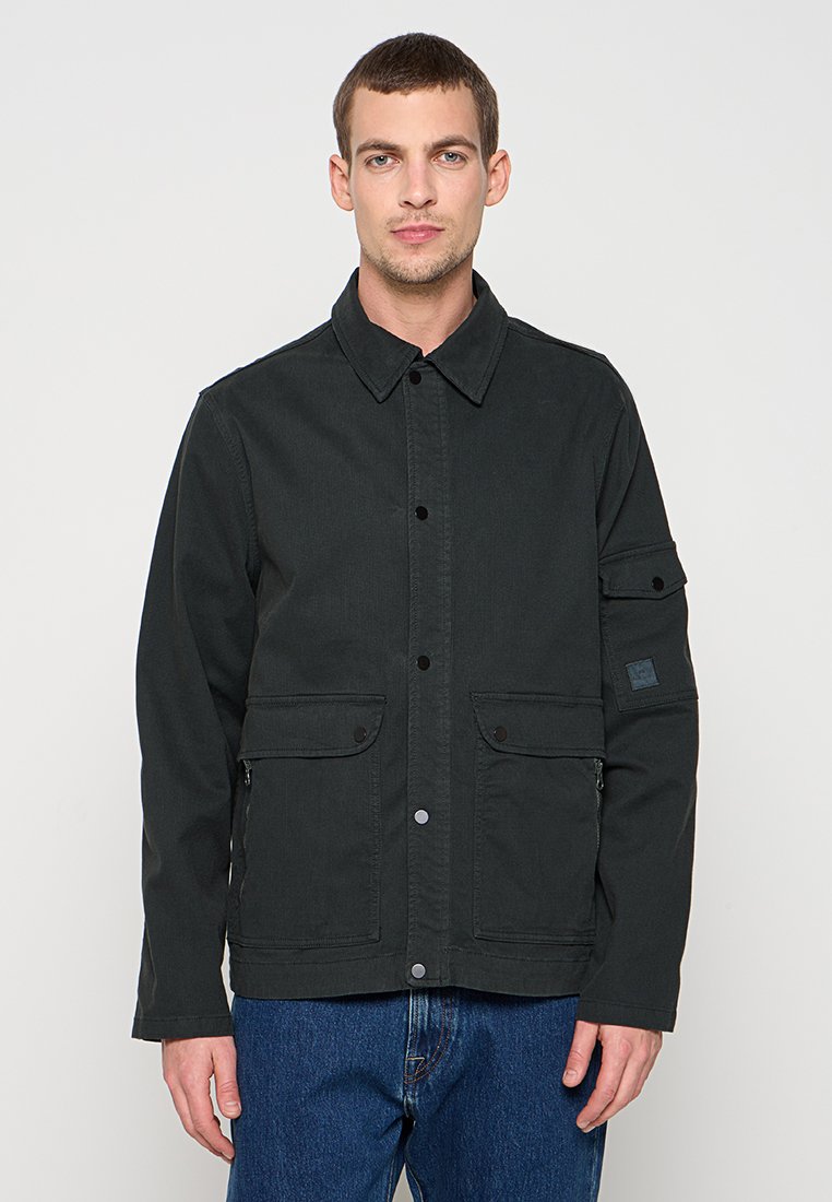 Schwarze Baumwolljacke mit Knopfleiste, zwei Fronttaschen und einer Reißverschlusstasche am Ärmel. Mit spitzem Kragen und lockerem Sitz.