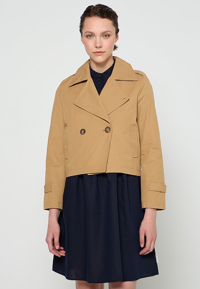 Beige Trenchcoat mit großen Revers, doppelreihigen Knöpfen und Seitentaschen, getragen über einem marineblauen Kleid. Glatte Textur.