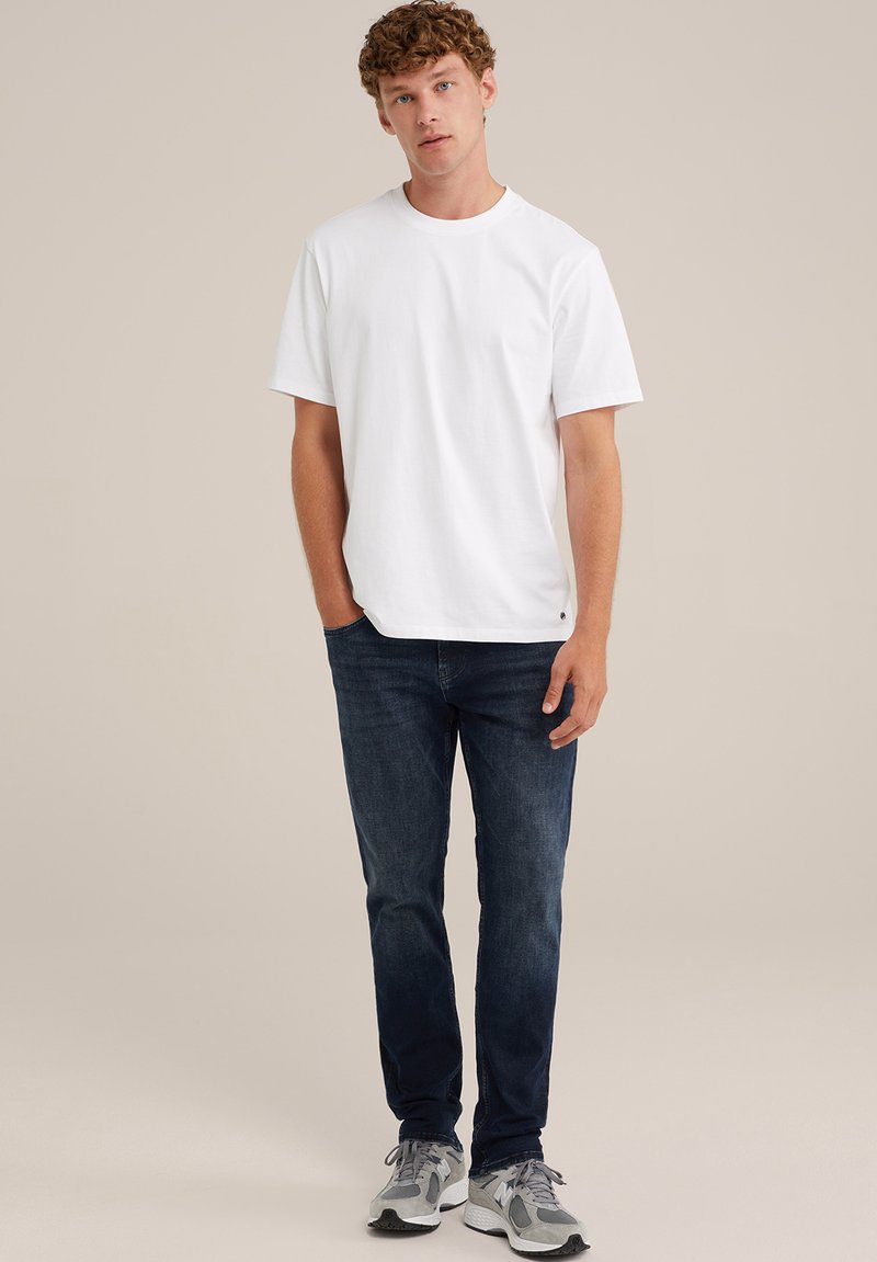 Weißes Baumwoll-T-Shirt mit rundem Ausschnitt, kombiniert mit dunkelblauen Jeans und grauen Sportschuhen mit dunkleren Akzenten.