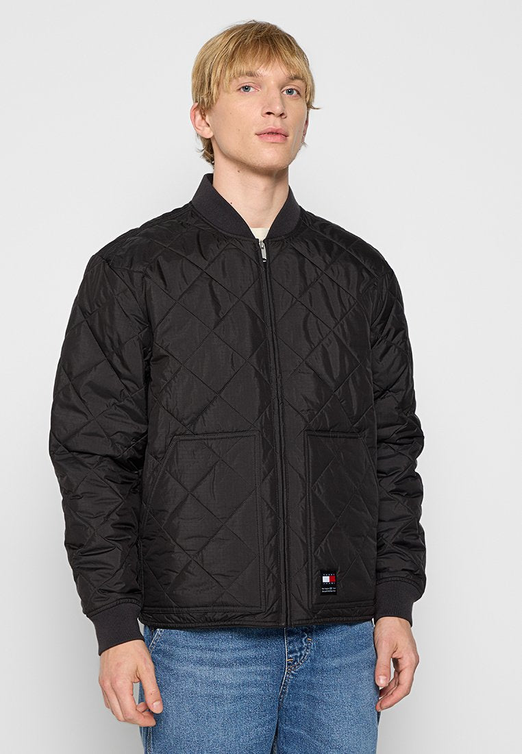 Junger Mann in einer schwarzen gesteppte Zip-Jacke mit gerippten Bündchen und Kragen, kombiniert mit blauen Jeans, vor einem schlichten hellen Hintergrund.