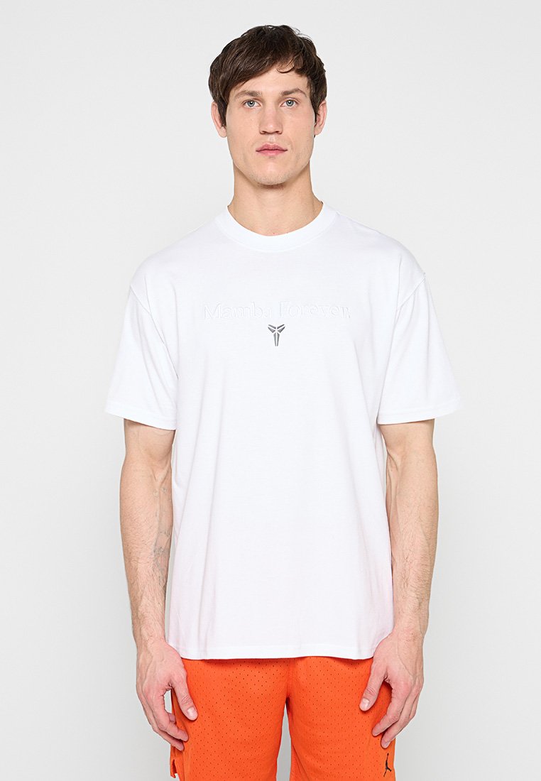 Junger Mann in weißem "Mamba Forever" T-Shirt und orangefarbenen Shorts steht vor hell einfarbigem Hintergrund.