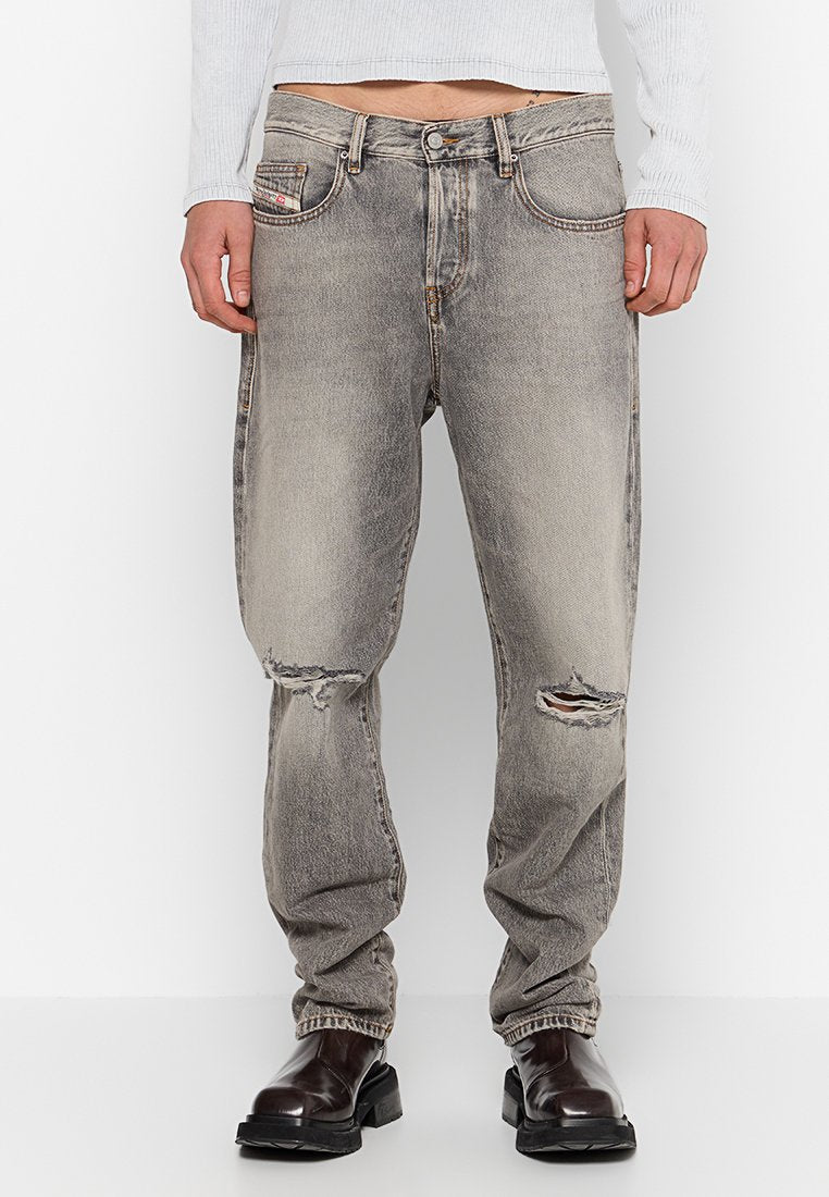 Person trägt ausgebleichte graue zerrissene Jeans mit Löchern an beiden Knien, ein weißes, geripptes Langarmoberteil und schwarze Lederstiefel.