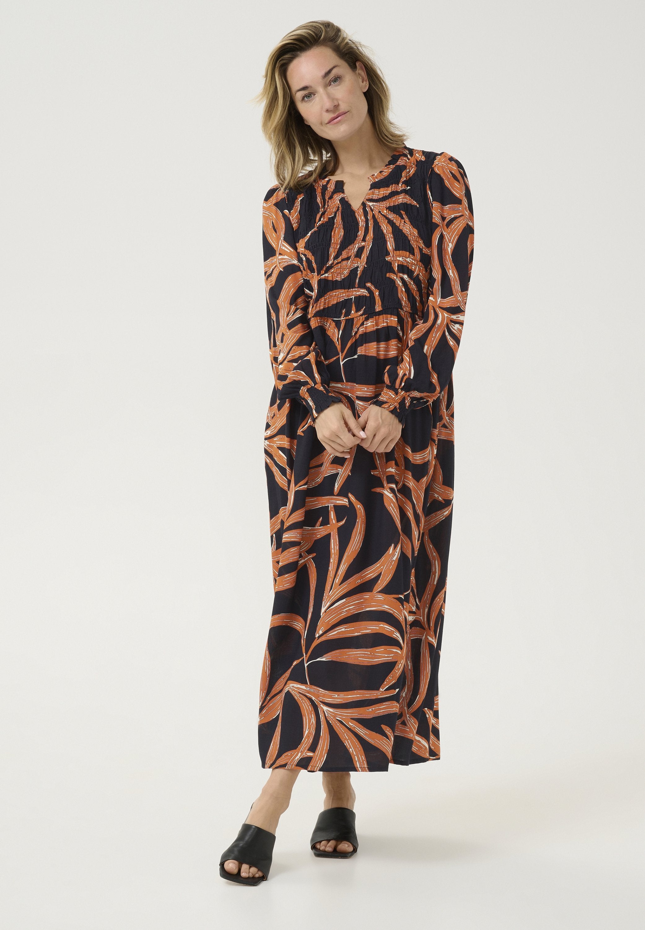 Langärmliges, marineblaues Kleid mit orangefarbenen Blätternmuster. Leichter Stoff, geraffter Oberteil und knöchellang. Kombiniert mit schwarzen Plateausandalen.