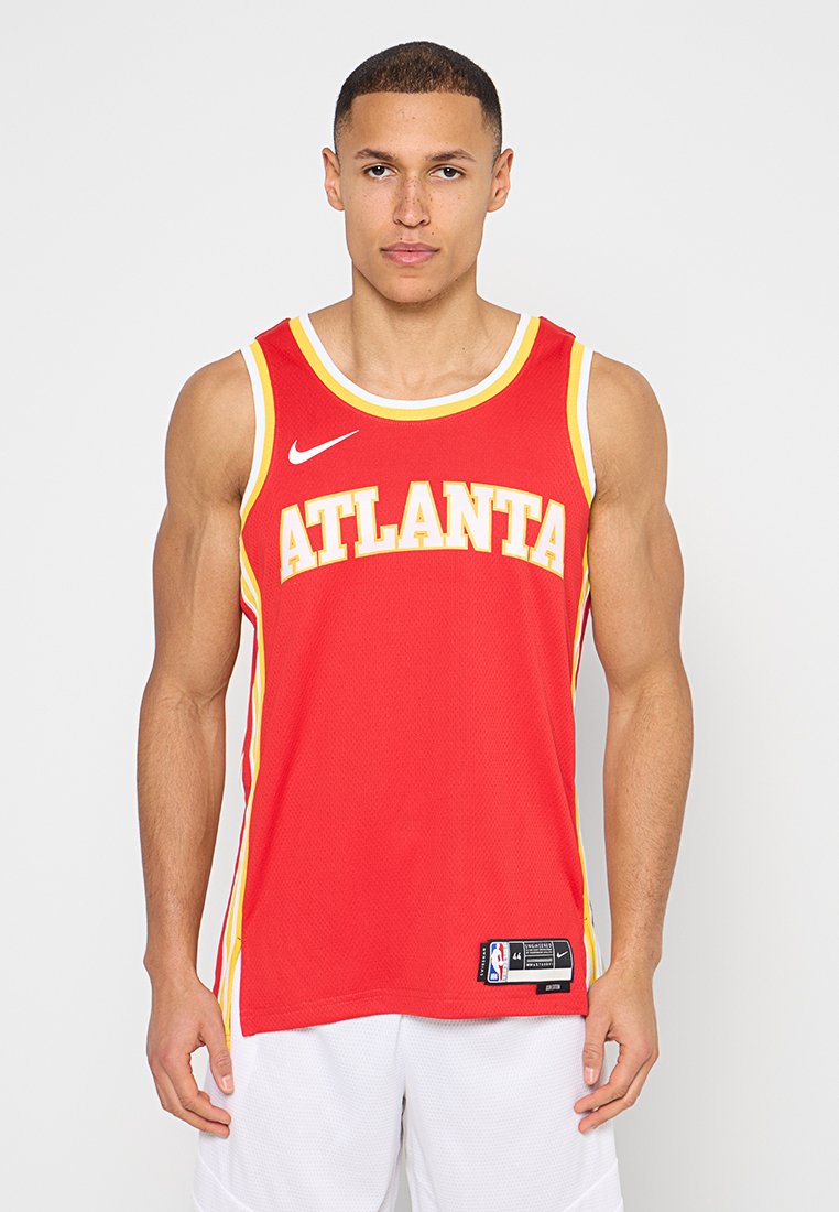 Junger Mann trägt ein rotes Atlanta-Basketballtrikot und weiße Shorts, steht vor einem schlichten weißen Hintergrund und schaut nach vorne.