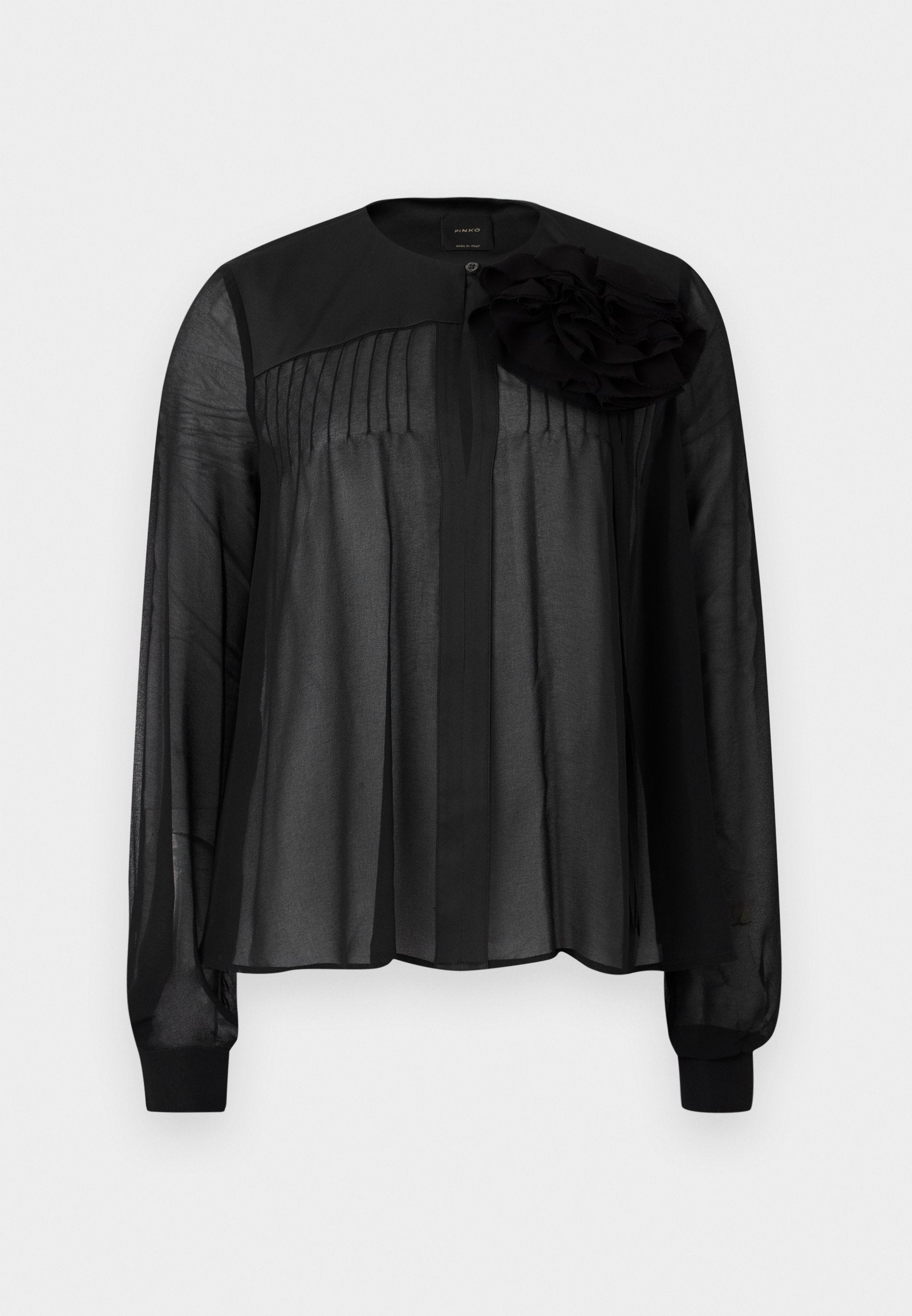 Schwarze Bluse mit langen transparenten Ärmeln, die vordere Falten und ein mehrlagiges Stoffblütenakzent an der linken Schulter aufweist. Knopfverschluss am Hals.