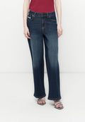 Person in dunkelblauen, weiten Jeans, einem roten Oberteil und offenen Sandalen mit Strassriemen, der vor einem schlichten, hellen Hintergrund steht.