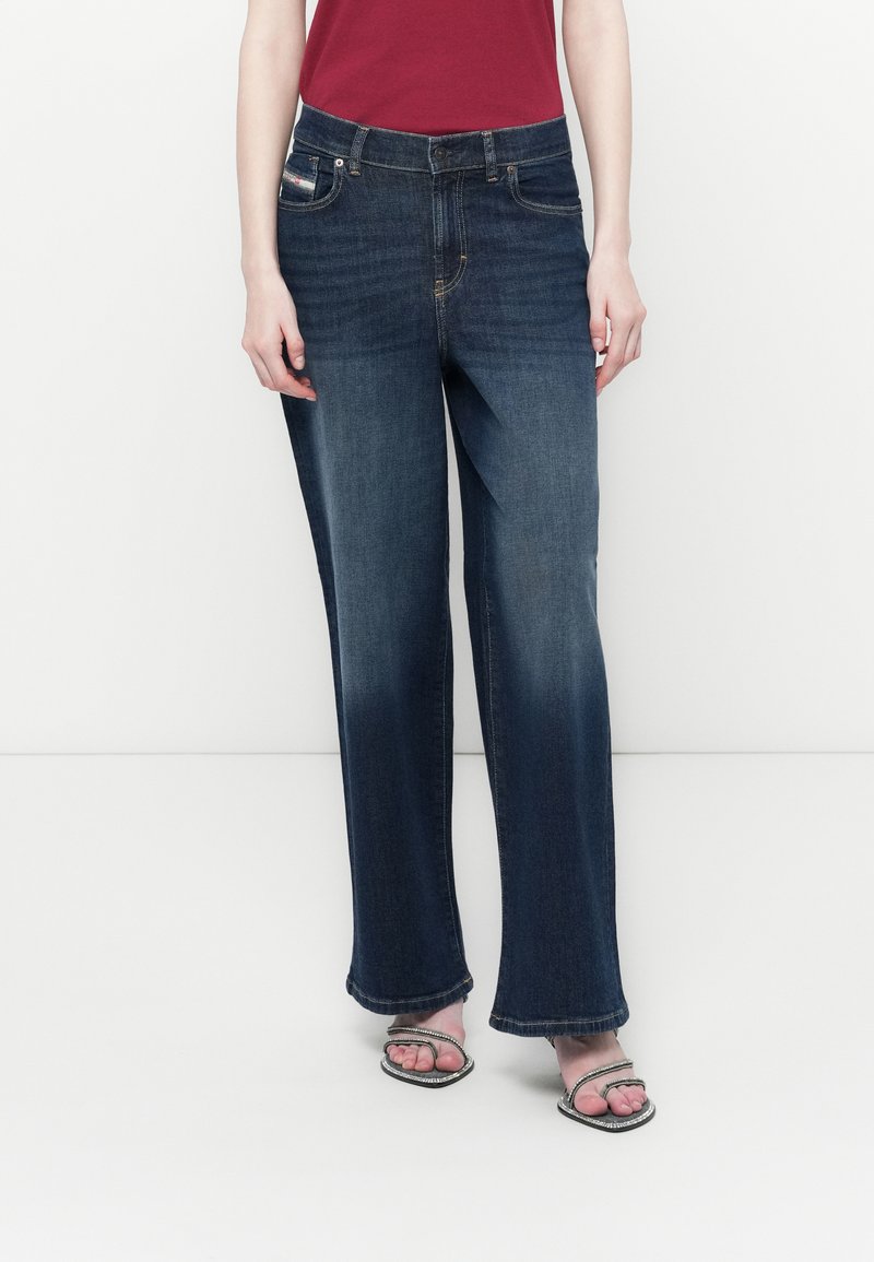 Person in dunkelblauen, weiten Jeans, einem roten Oberteil und offenen Sandalen mit Strassriemen, der vor einem schlichten, hellen Hintergrund steht.