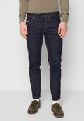 Dunkelblaue Slim-Fit-Jeans aus Denim mit sichtbaren Nähten, fünf Taschen und einem vorderen Reißverschluss, kombiniert mit grünen Schuhen und einem beigen Oberteil.