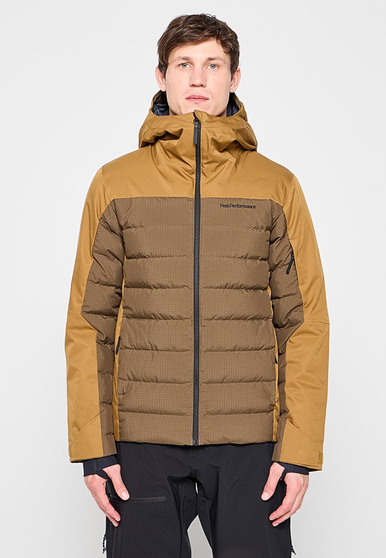 Mann trägt braune zweifarbige gesteppte Jacke mit Kapuze, hochgezogen, steht vor einfarbigem hellem Hintergrund mit entspannt an den Seiten hängenden Händen.