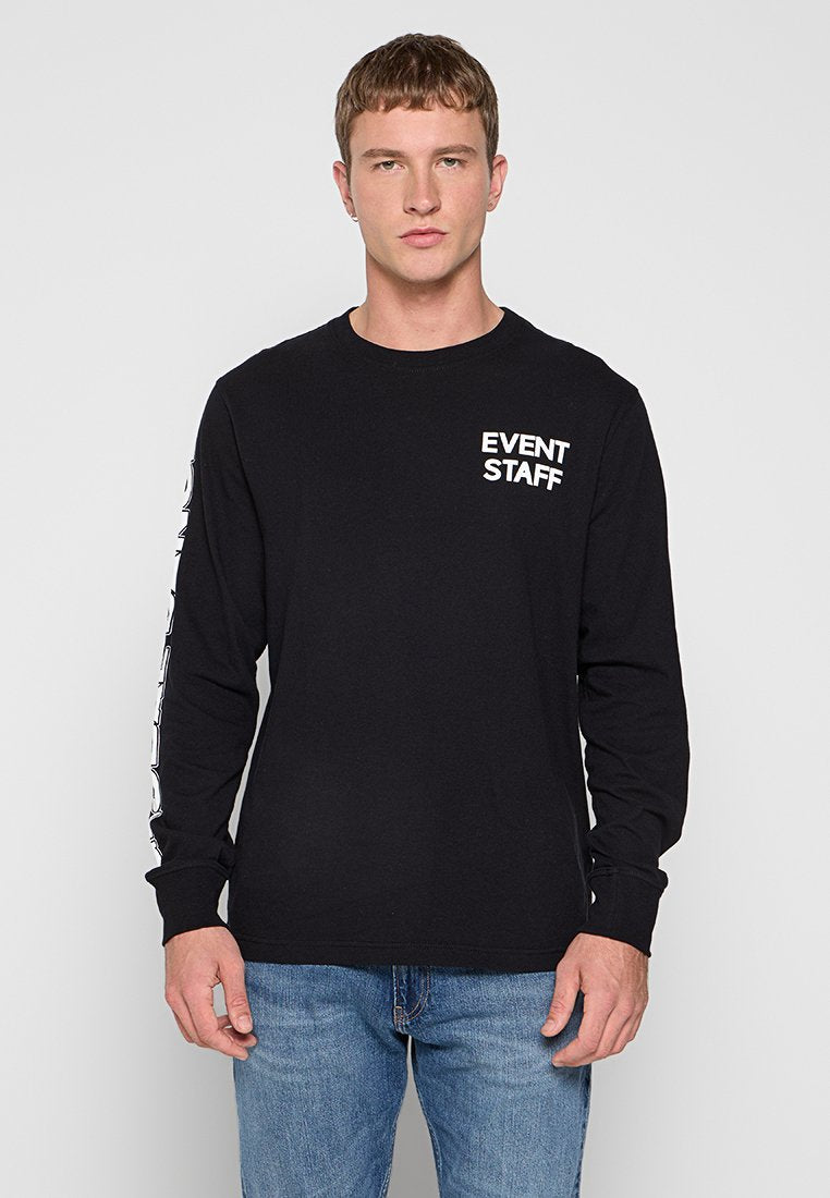 Mann in schwarzem Langarmshirt mit dem Text "EVENT STAFF" auf der Brust und dem Ärmel, kombiniert mit blauen Jeans, steht vor einem neutralen Hintergrund.