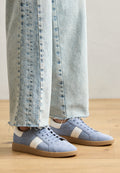 Helles blaugraues Wildleder-Sneaker mit weißen Streifen, runder Spitze und Schnürdesign. Getragen mit ausgefransten hellen Jeans auf einem Holzboden.