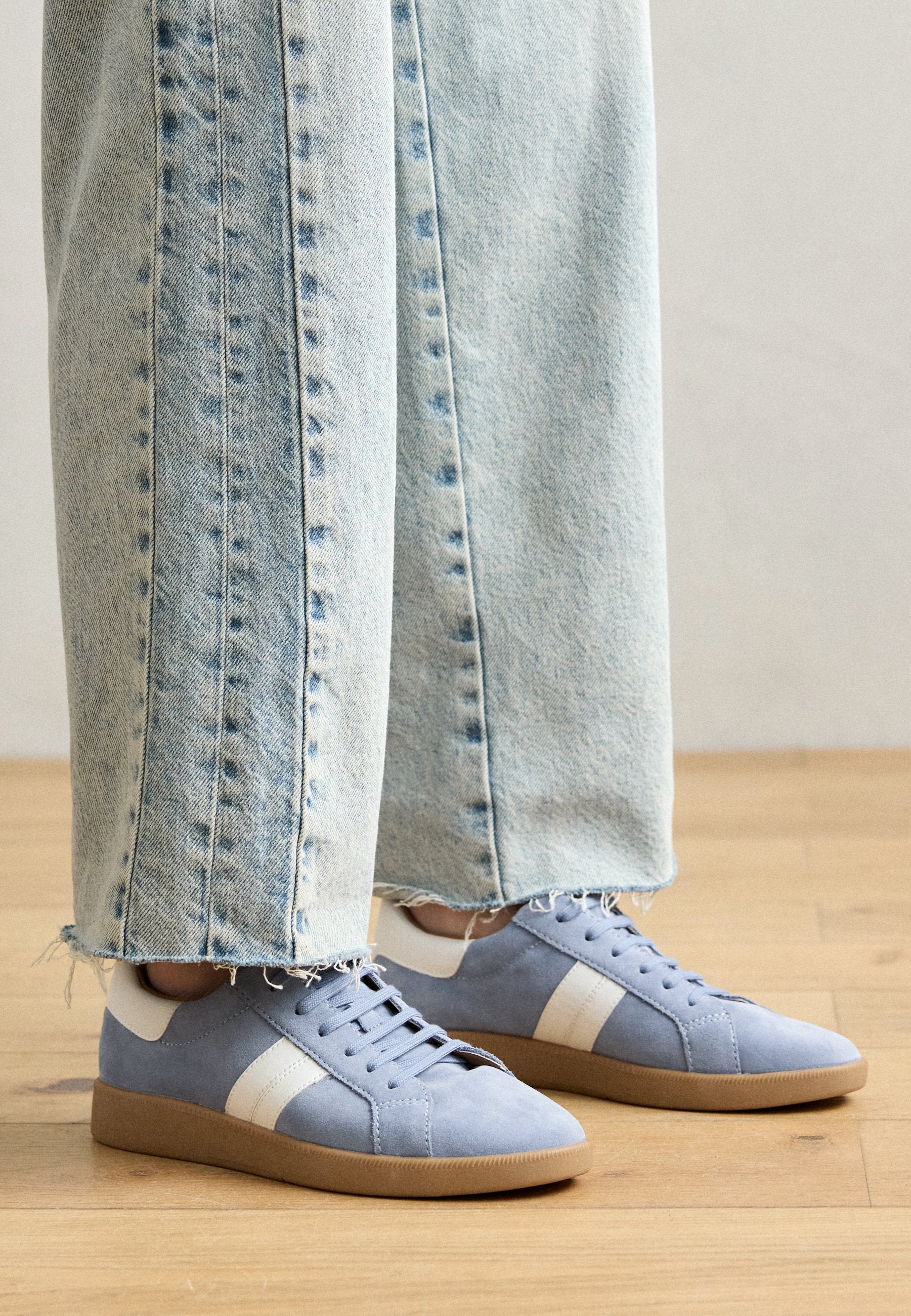 Helles blaugraues Wildleder-Sneaker mit weißen Streifen, runder Spitze und Schnürdesign. Getragen mit ausgefransten hellen Jeans auf einem Holzboden.