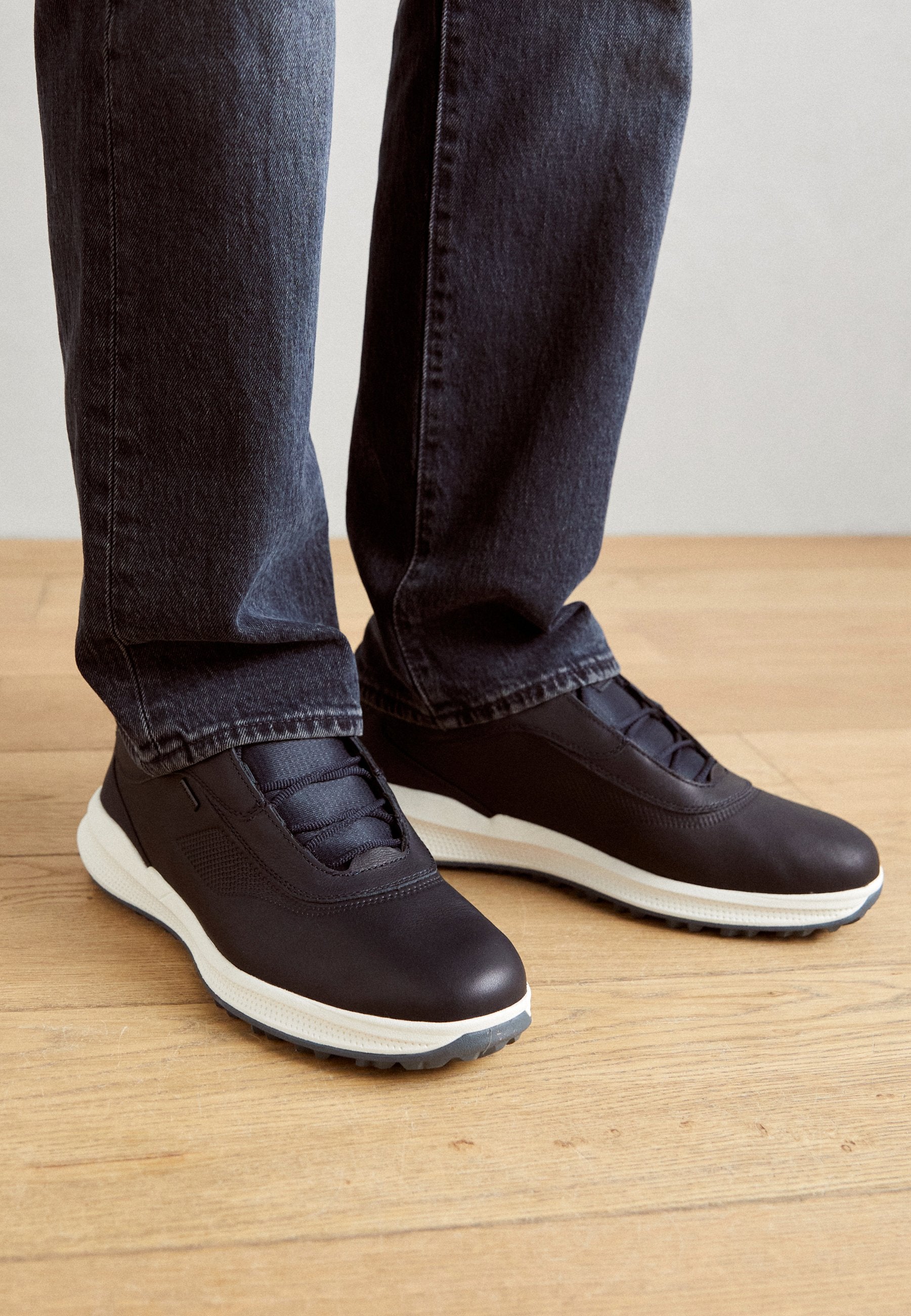 Schwarze, niedrige Sneakers mit synthetischem Obermaterial, strukturierten Akzenten und weißer Zwischensohle. Getragen mit dunklen Denim-Jeans auf einem Holzboden.
