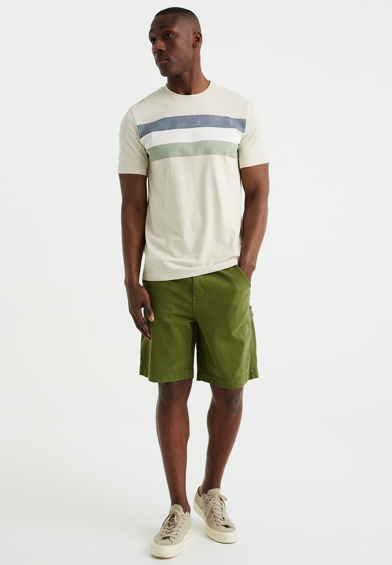 Beige T-Shirt für Herren mit grünen, blauen und weißen Streifen, kombiniert mit grünen Shorts. Aus Baumwollmaterial, einfaches Design, lässiger Schnitt. Beige Sneaker.