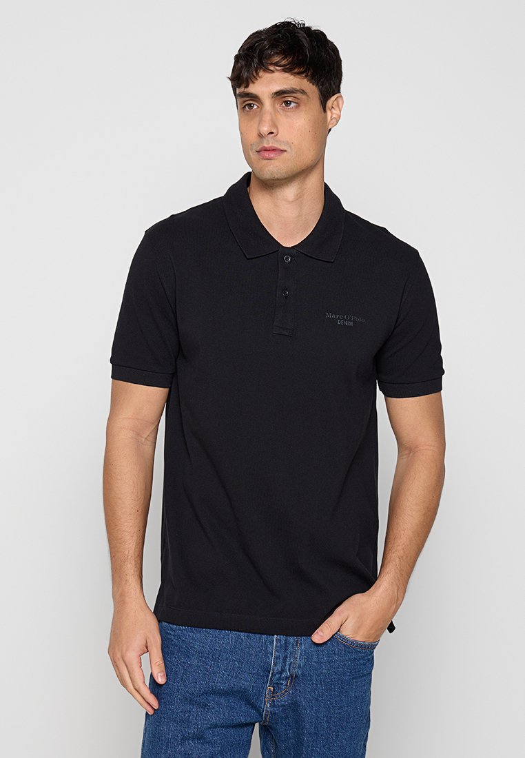 Mann trägt schwarzes kurzärmeliges Poloshirt und blaue Jeans, steht mit einer Hand in der Tasche vor einem schlichten hellgrauen Hintergrund.