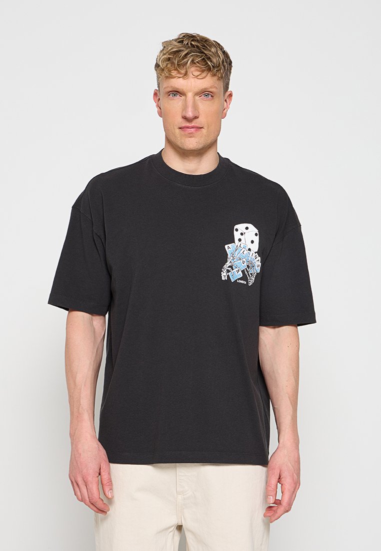 Mann trägt ein schwarzes T-Shirt mit Würfeln und blauen Spielkarten-Grafiken auf der Brust, kombiniert mit hellen Hosen.