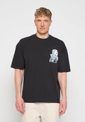 Mann trägt ein schwarzes T-Shirt mit Würfeln und blauen Spielkarten-Grafiken auf der Brust, kombiniert mit hellen Hosen.