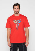 Mann mit kurzen lockigen Haaren und Bart, der ein rotes T-Shirt mit einem bunten geometrischen Schmetterlingslogo auf der Vorderseite trägt.